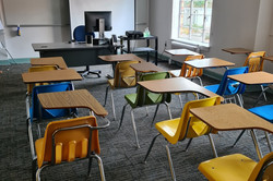 Hier sieht man ein Klassenzimmer mit zahlreichen Einzeltischen, an denen Stühle stehen. Vorne ist ein Whiteboard und ein Computer.